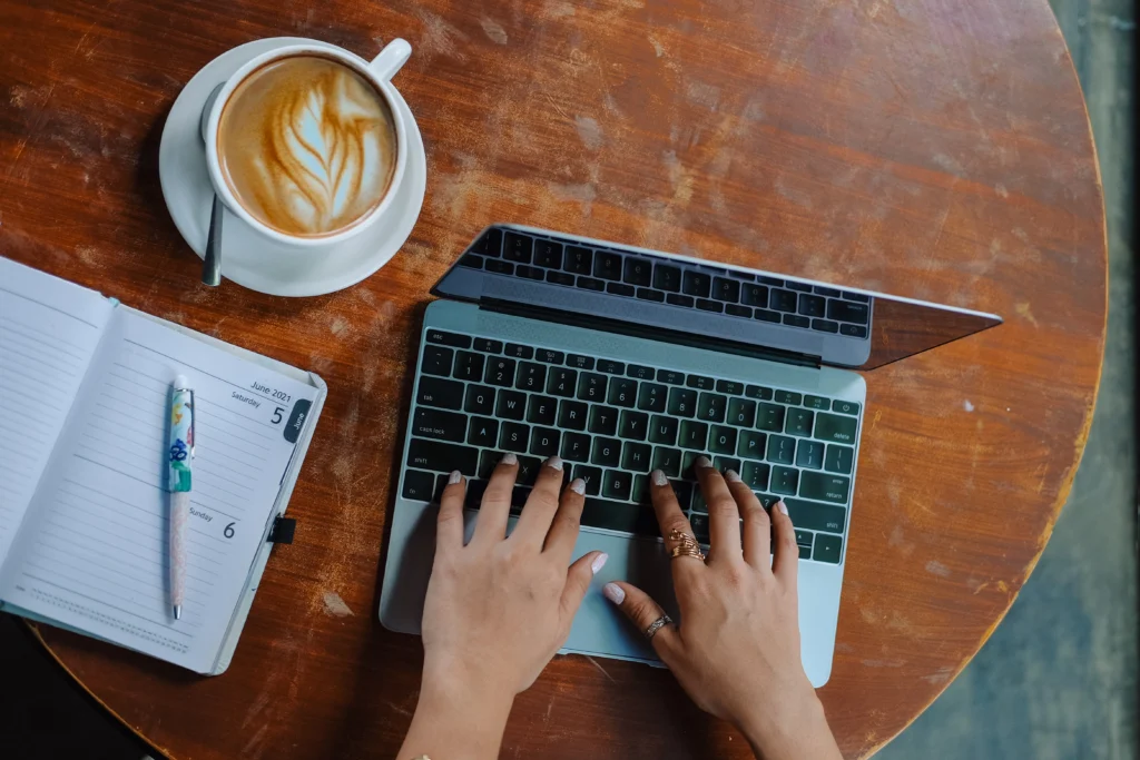 Hand typing on laptop with coffee and notebook on the side